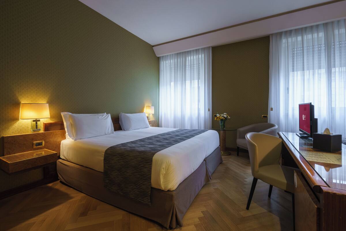 Une chambre d’hôtel avec un grand lit, un bureau avec un ordinateur, une chaise et une petite table sur laquelle est posé un vase de fleurs.