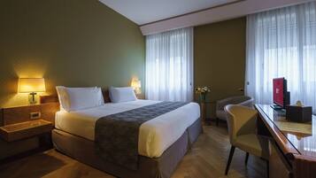 A hotel room with a large bed, a desk with a computer, a chair, and a small table with a vase of flowers.