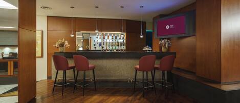 A bar area with a wooden bar counter, three red bar stools, a wall-mounted TV, and a shelf with bottles.