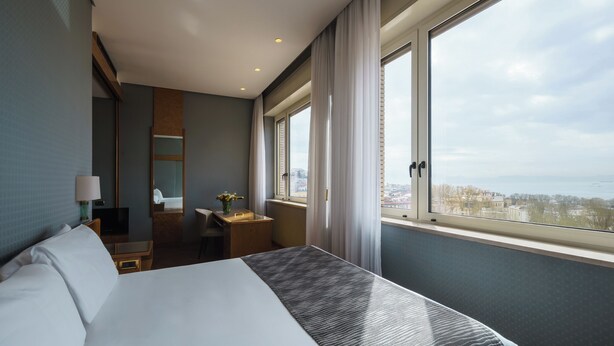 A hotel room with a large bed, a desk, a chair, a TV, and a window with a city view. - Exe Majestic (Naples)
