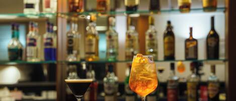 Two cocktails on a bar counter with a background of various liquor bottles on shelves.