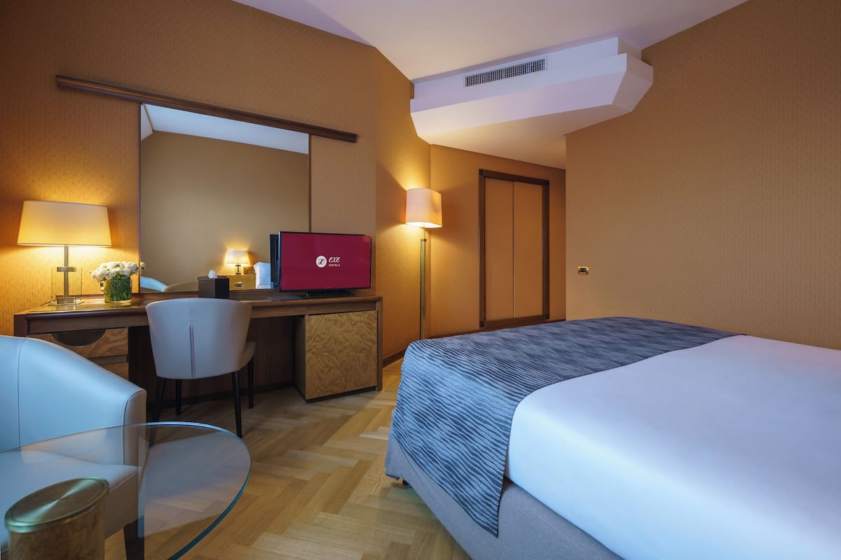 A hotel room with a bed, a desk with a computer, a chair, and a lamp.