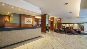 A modern hotel lobby with a reception desk, seating area, and decorative plants. - Exe Majestic (Naples)