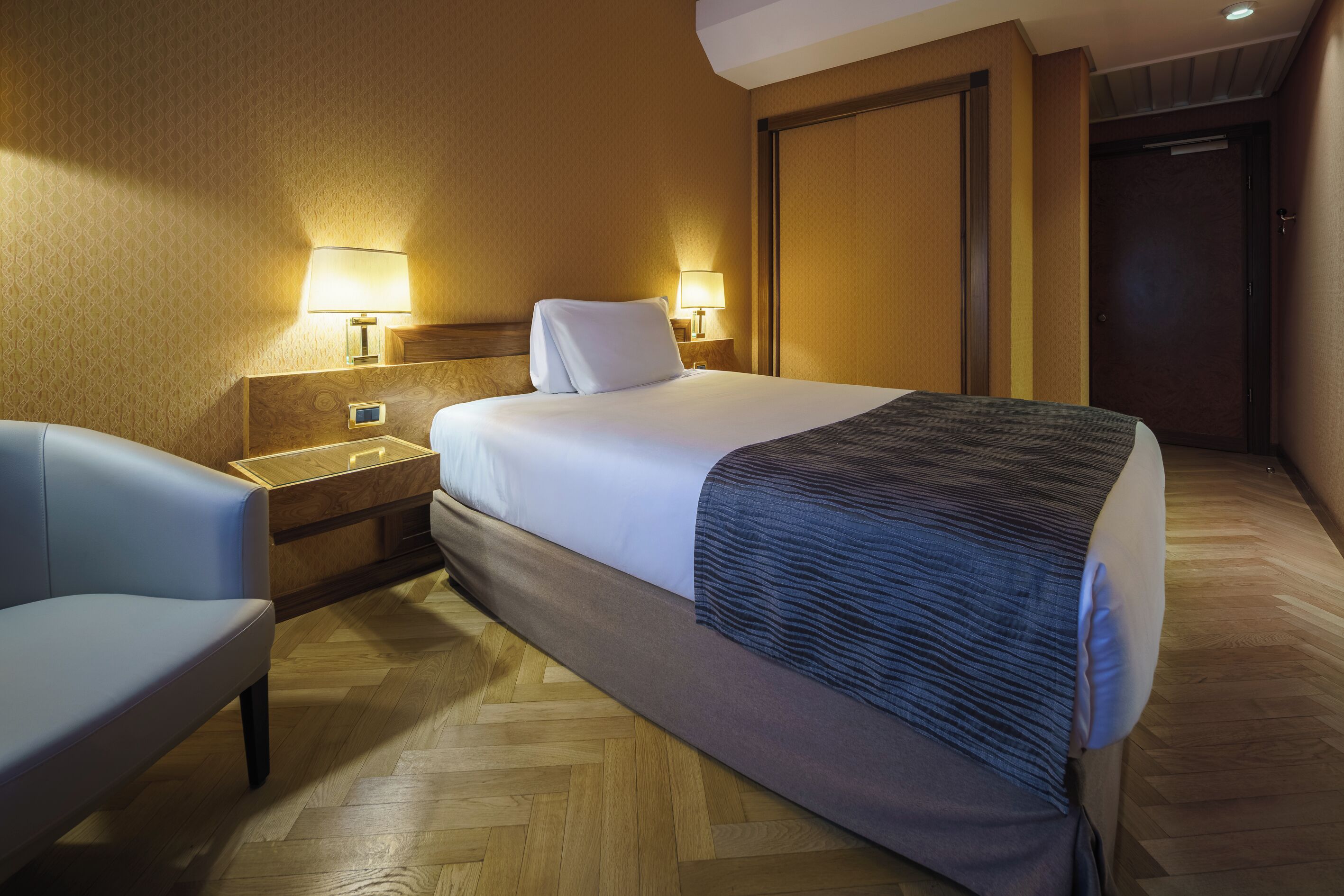 A hotel room with a bed, bedside tables, a chair, and a wooden floor.