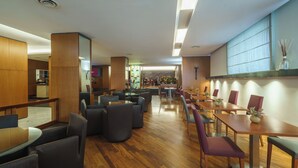 A modern hotel lobby with wooden flooring, leather seating, and a mix of round and rectangular tables. - Exe Majestic (Naples)