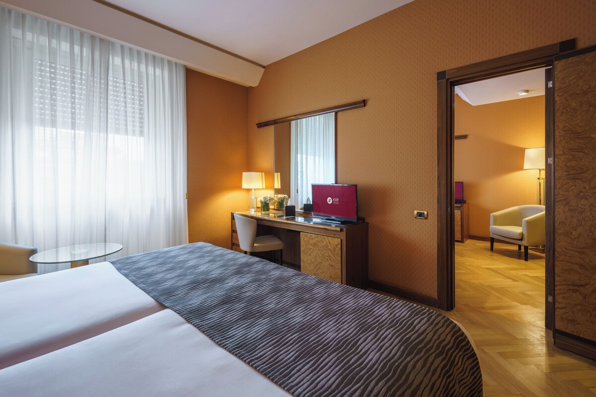 Una habitación de hotel con una cama grande, un escritorio con una computadora portátil, una silla, una mesa redonda y una puerta que da a otra habitación.