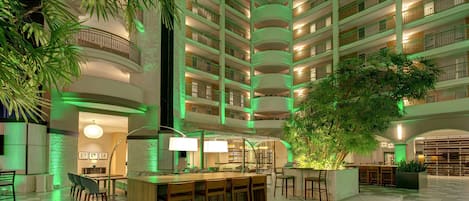 A multi-level hotel lobby with a central bar area, multiple balconies, and indoor plants.