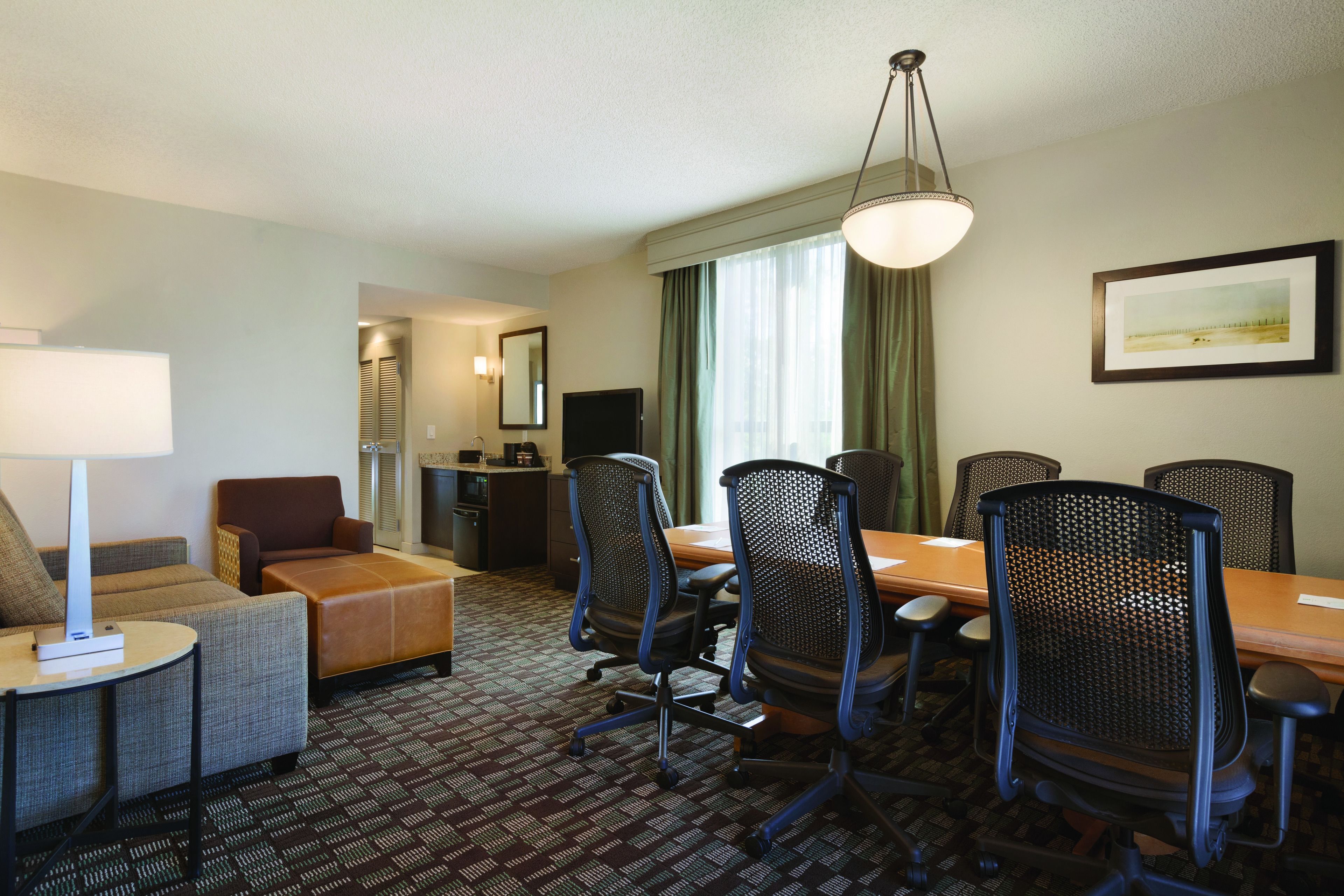 A hotel room with a dining area, a sofa, a desk with a lamp, and a television.
