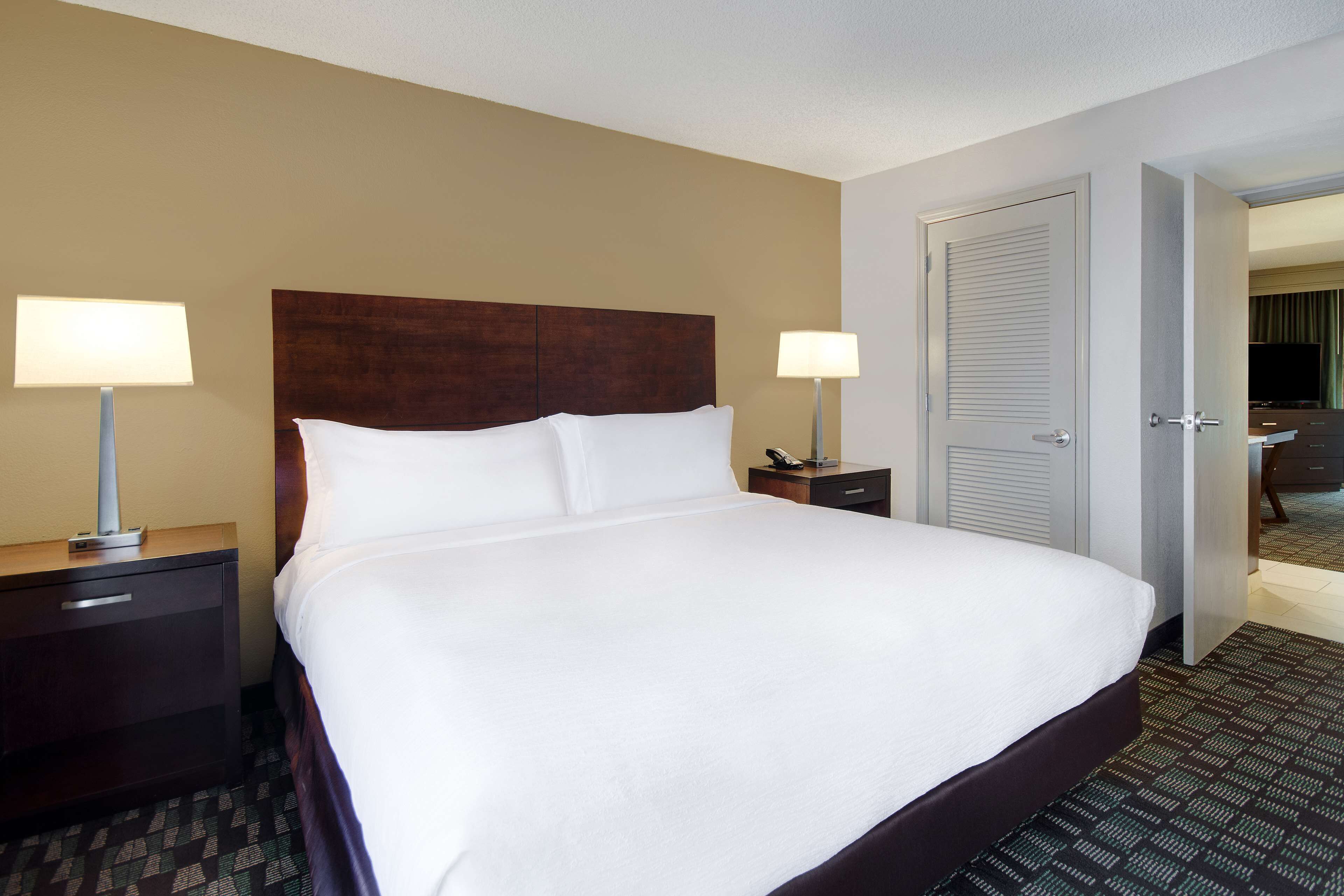 A hotel room with a large bed, two bedside tables with lamps, a wooden headboard, and a door leading to another room.