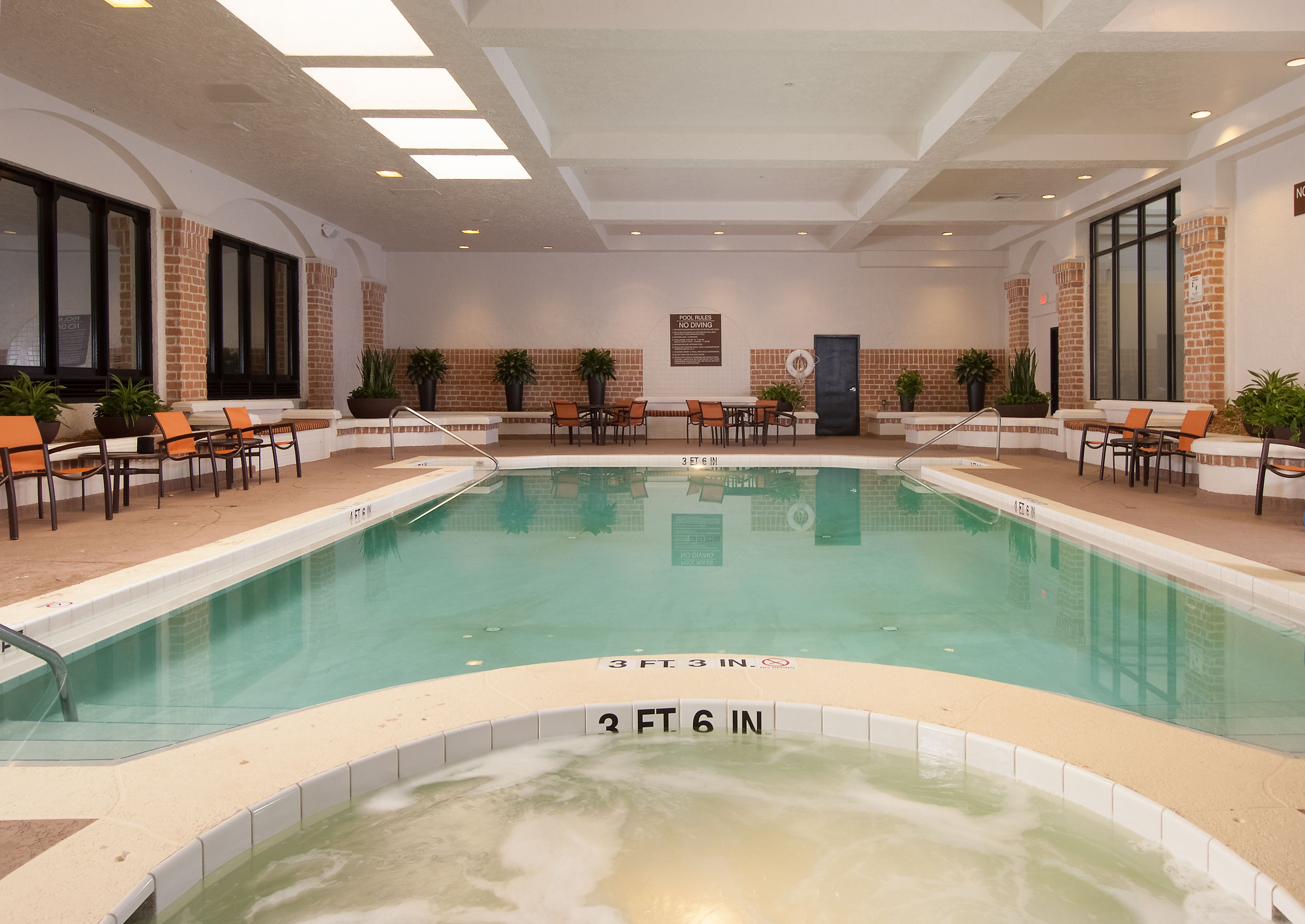 Una piscina interior con una profundidad de 3 pies y 6 pulgadas, rodeada de sillas de descanso y mesas, con paredes de ladrillo y ventanas amplias.