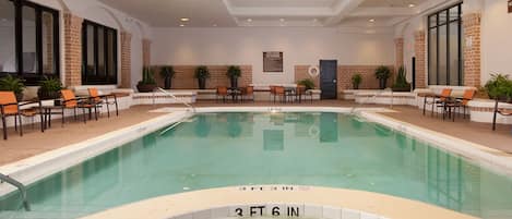 Una piscina interior con una profundidad de 3 pies y 6 pulgadas, rodeada de sillas de descanso y mesas, con paredes de ladrillo y ventanas amplias.