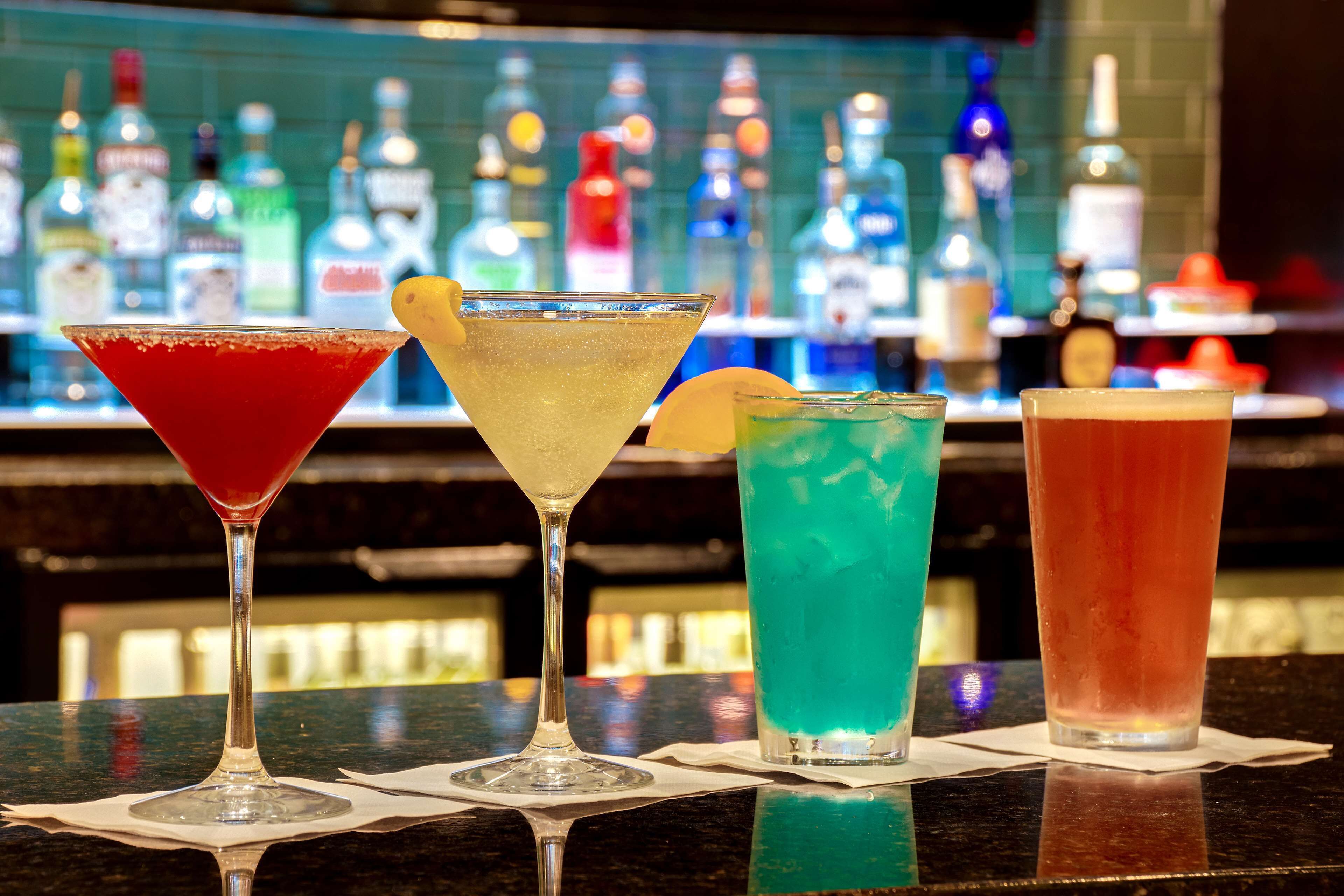 Four cocktails on a bar counter with a blurred background of various bottles.