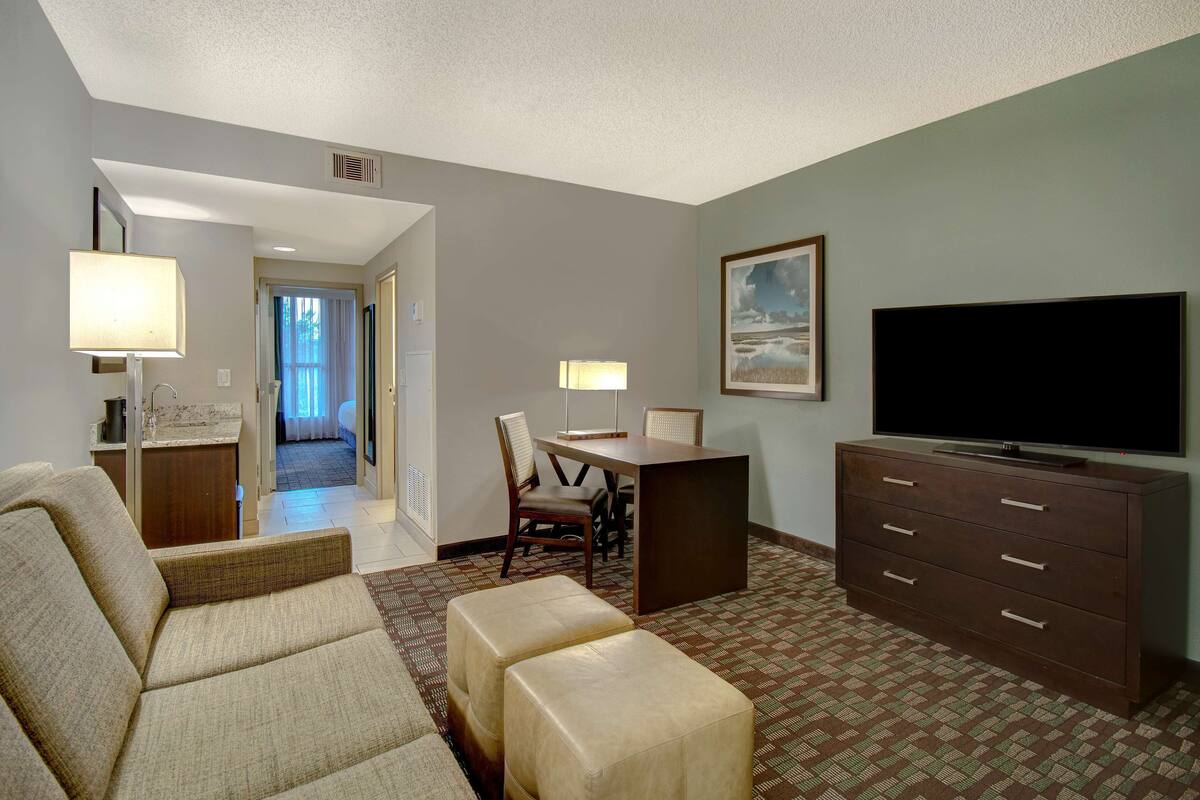Una habitación de hotel con sofá, reposapiés, mesa de comedor, sillas, televisor y cómoda.
