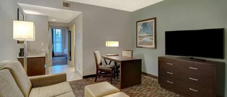 A hotel room with a sofa, ottoman, dining table, chairs, a TV, and a dresser.
