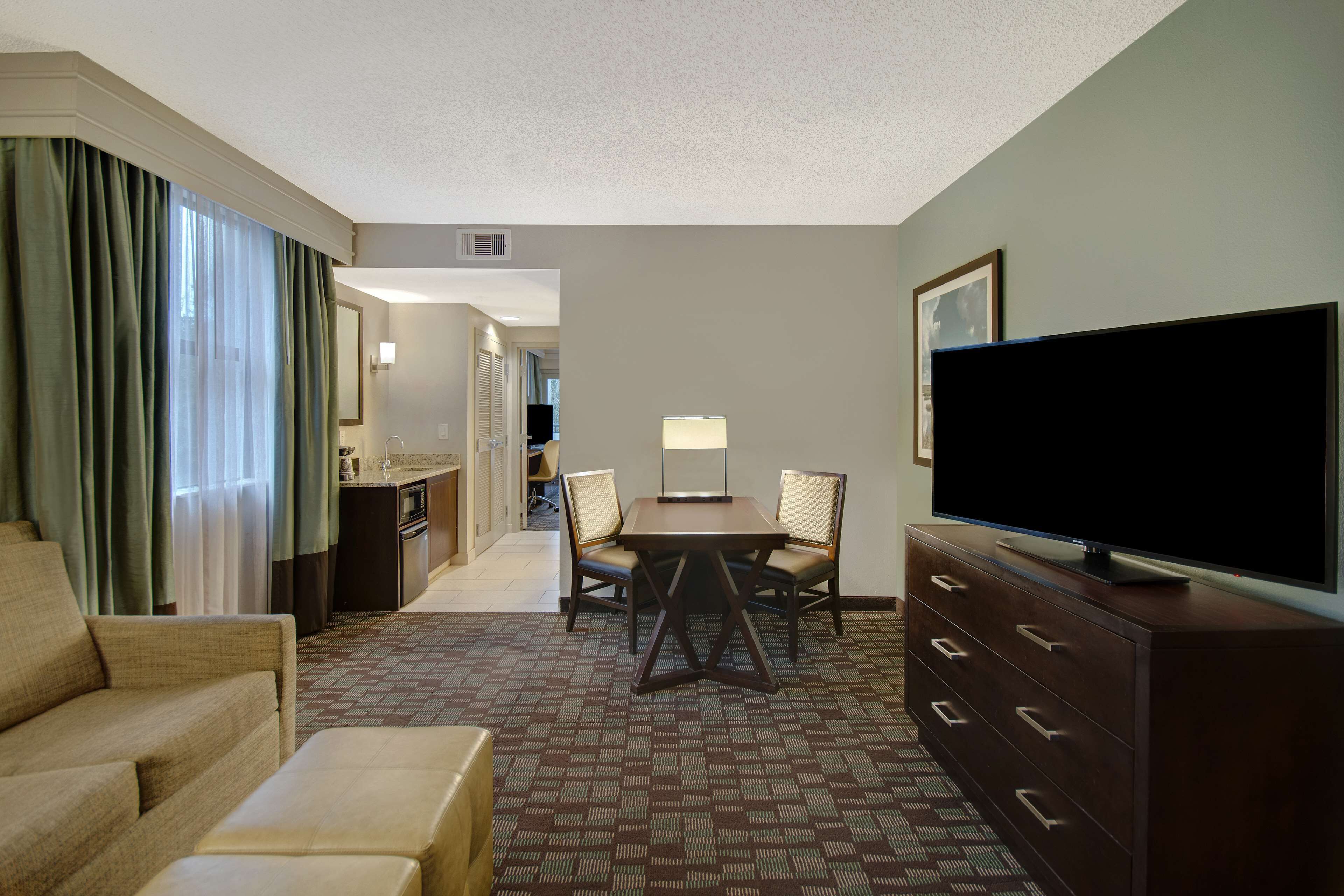 A hotel room with a sofa, a TV on a dresser, a dining table with chairs, and a kitchen area in the background.