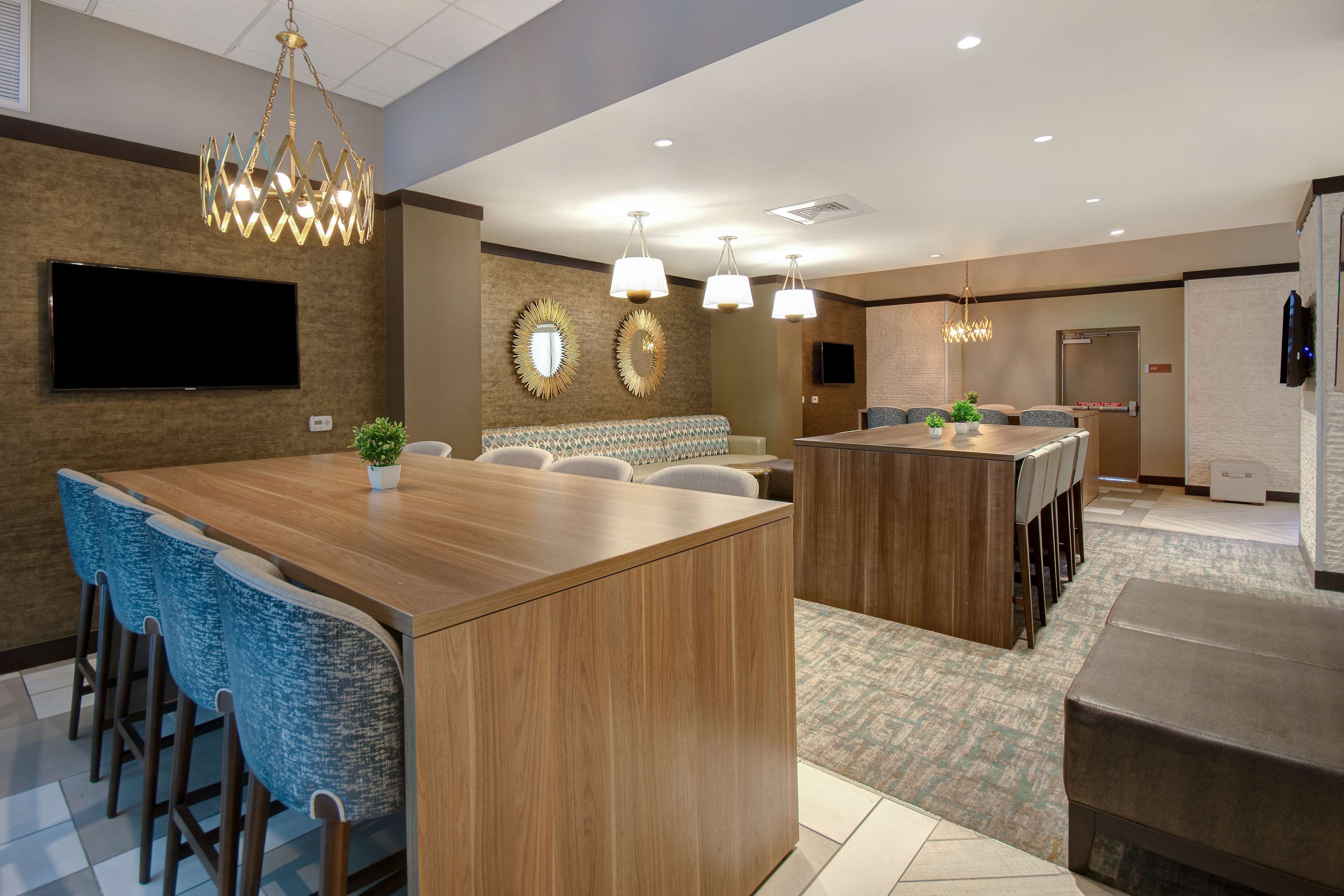 A modern hotel lobby with a central wooden reception desk, blue upholstered bar stools, a seating area with patterned sofas, and a chandelier.