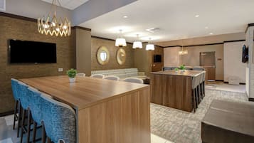 A modern hotel lobby with a central wooden reception desk, blue upholstered bar stools, a seating area with patterned sofas, and a chandelier.
