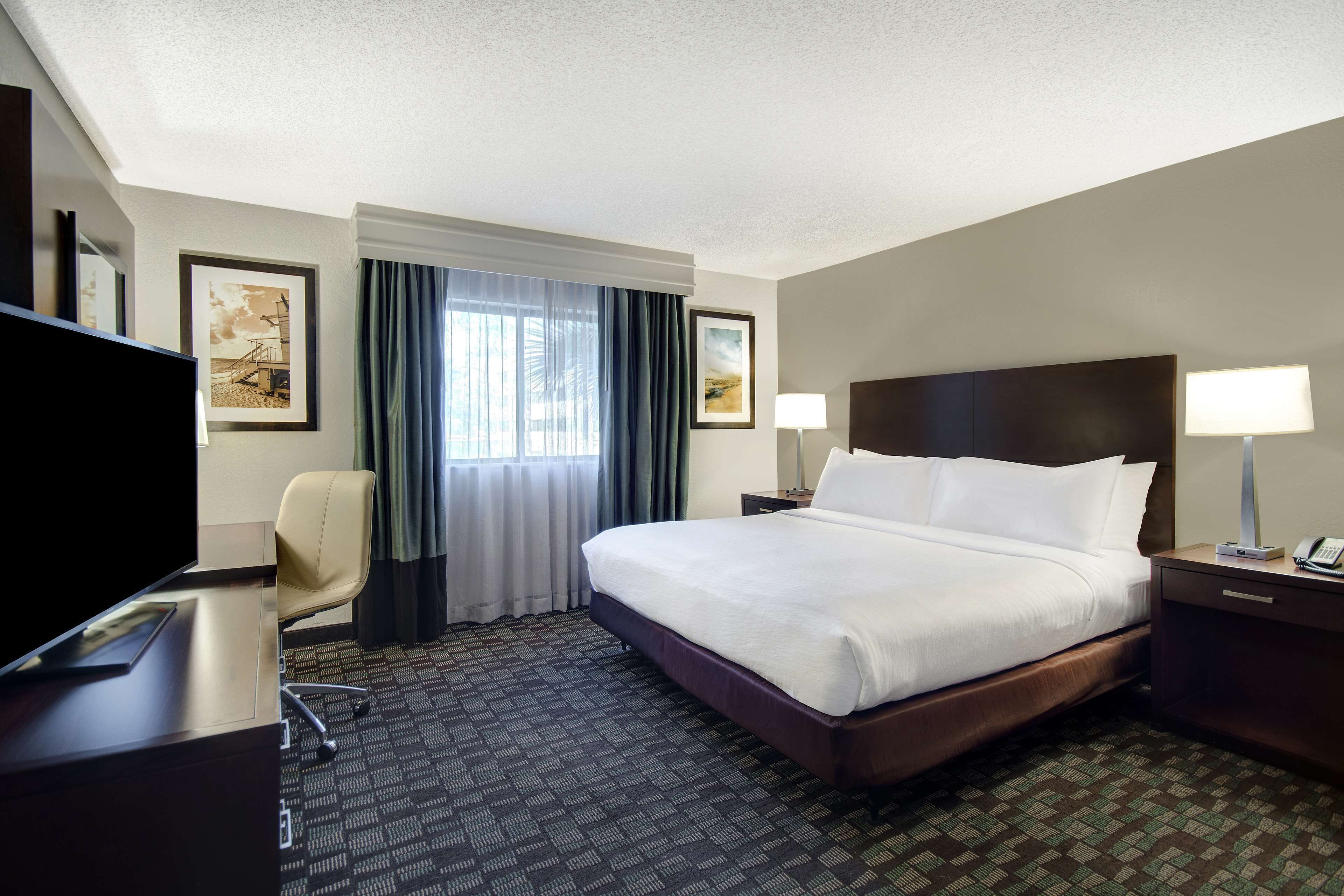 A hotel room with a large bed, a desk with a flat-screen TV, a chair, a nightstand with a lamp, and framed pictures on the wall.