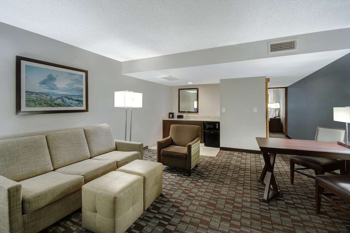 Habitación de hotel con sofá, sillón, reposapiés, mesa de comedor y una zona de kitchenette.