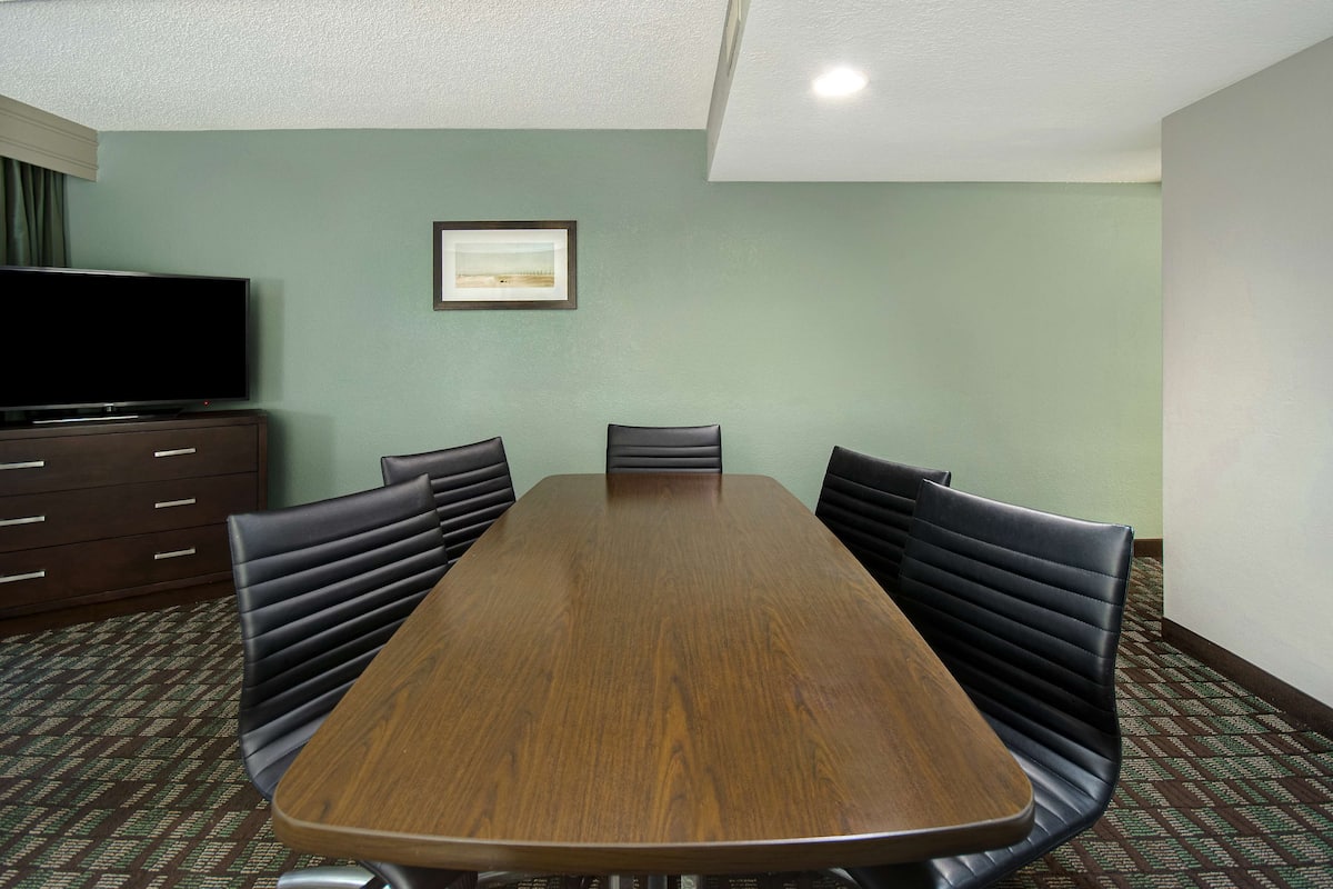 Un salón de conferencias con una mesa de madera, sillas tapizadas de color negro, un televisor sobre un mueble y un cuadro en la pared.
