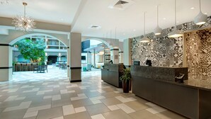 Un vestíbulo de hotel con mostrador de recepción, pared con diseño, lámparas colgantes, una planta grande y una amplia zona de estar con mesas y sillas.