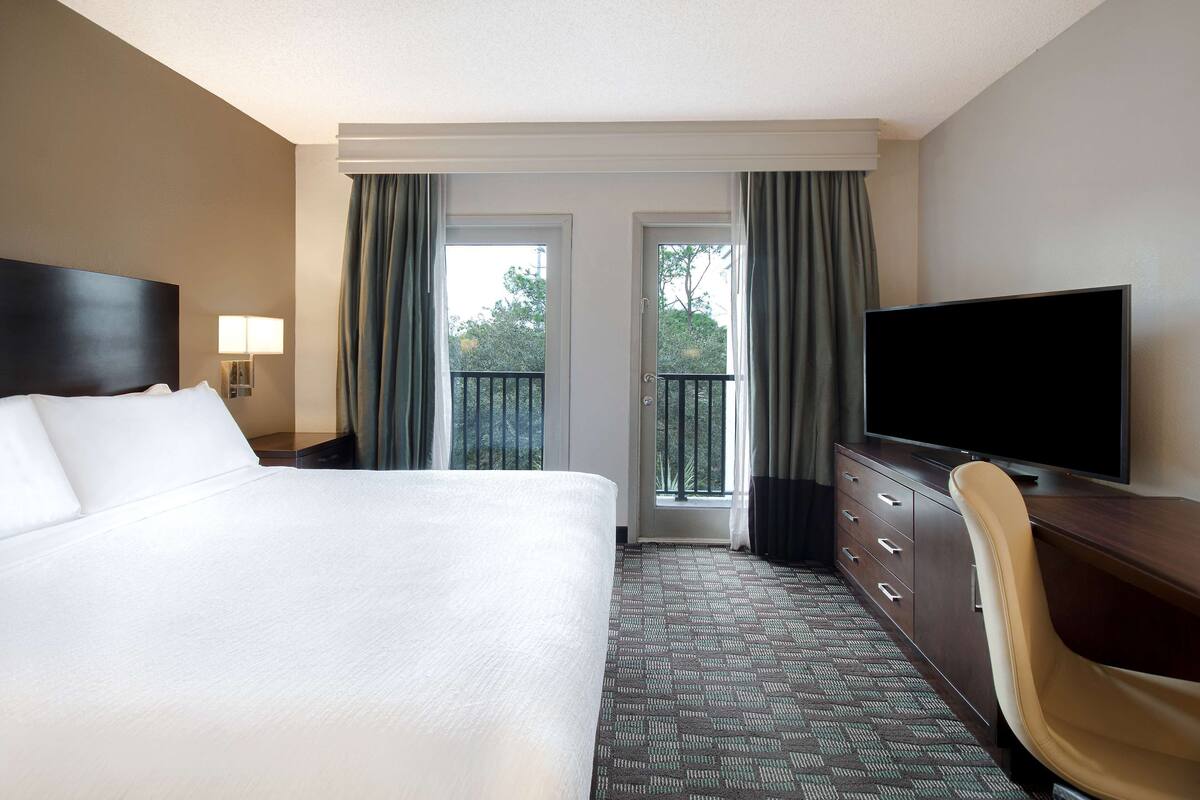 Habitación de hotel con una cama grande, televisor, escritorio y vistas al exterior a través de una puerta.