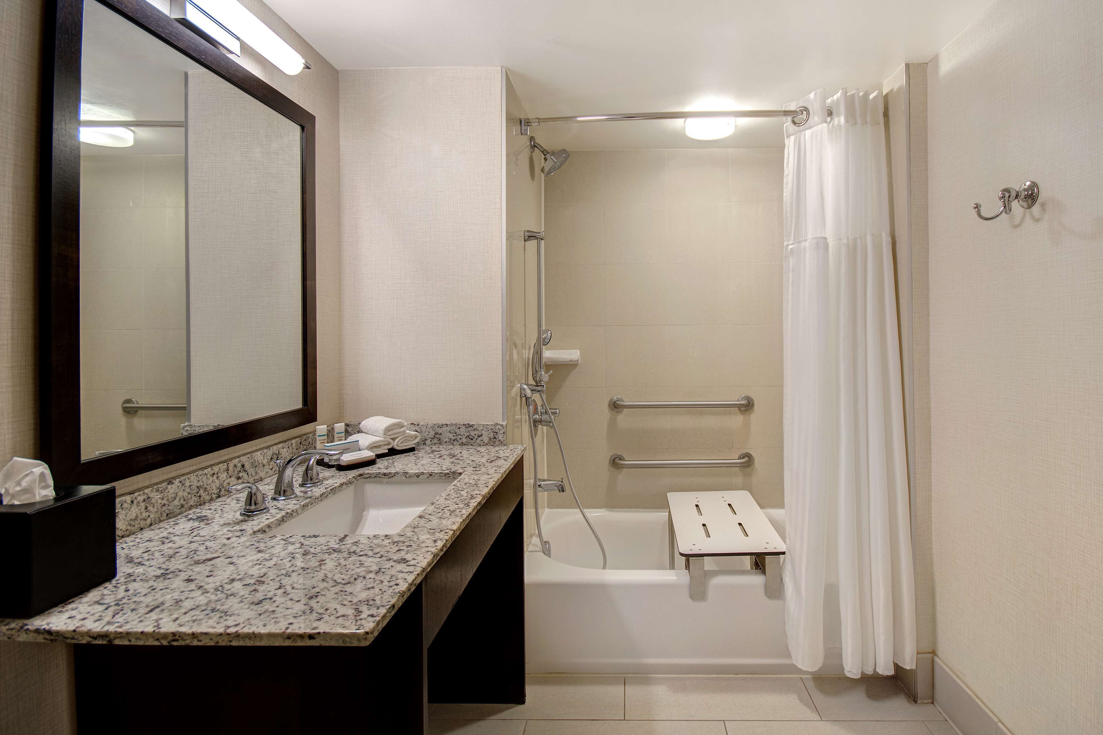 A bathroom with a granite countertop sink, a mirror, a shower with a handheld showerhead, and a white shower curtain.