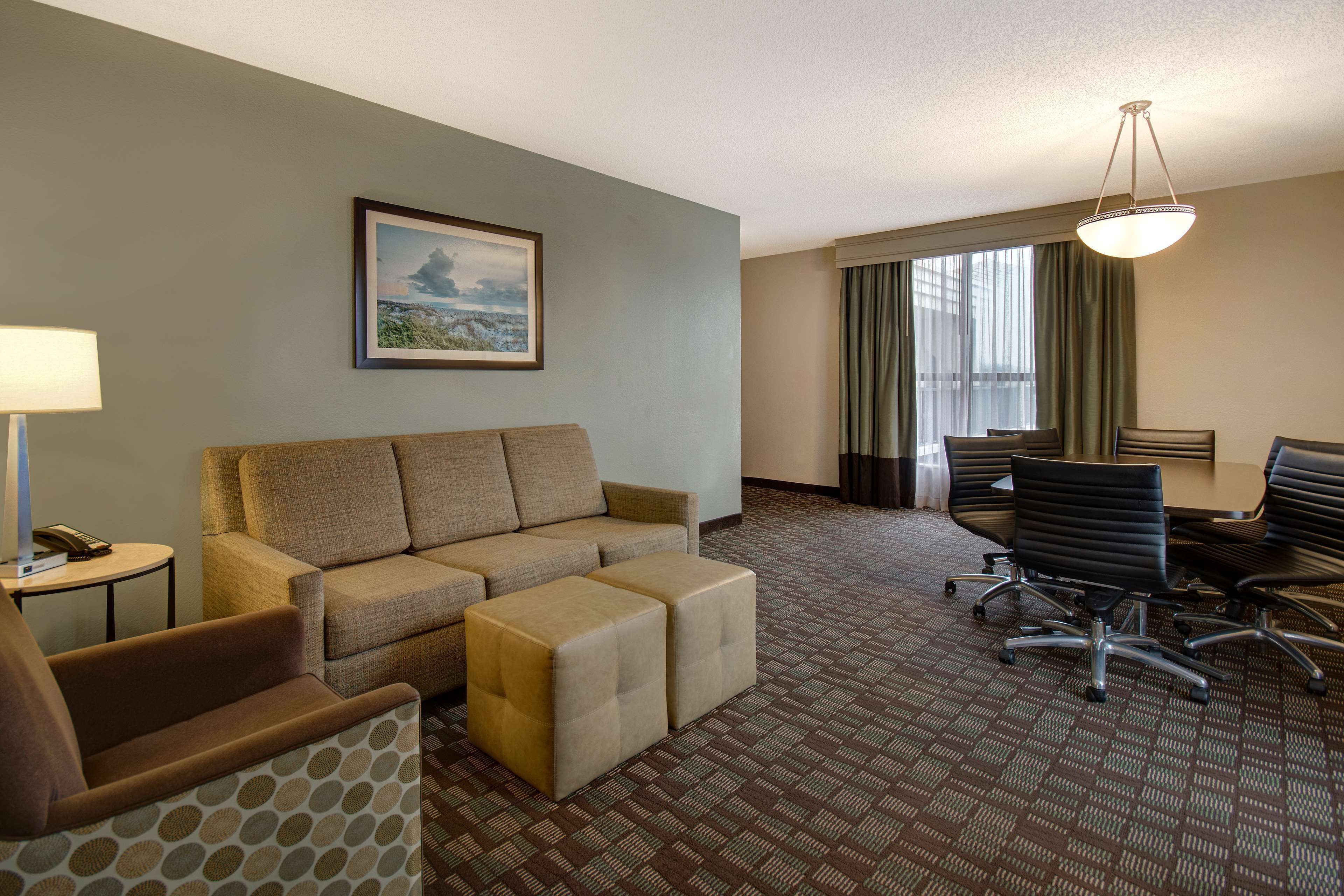 A hotel room with a sofa, ottoman, a small table with a lamp, a painting on the wall, a dining table with chairs, and a patterned carpet.
