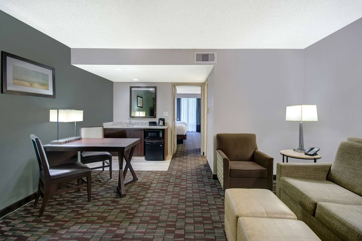 Habitación de hotel con comedor, sofá, una mesita con lámpara y cama al fondo.