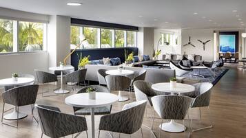 Un moderno vestíbulo de hotel con mesas redondas, sillas tapizadas en gris, un área de sofás y grandes ventanales con vista a palmeras.
