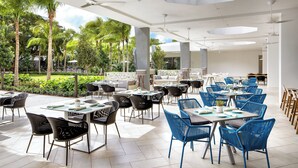 Amplia zona de comedor al aire libre con mesas y sillas, rodeada de exuberante follaje y palmeras.