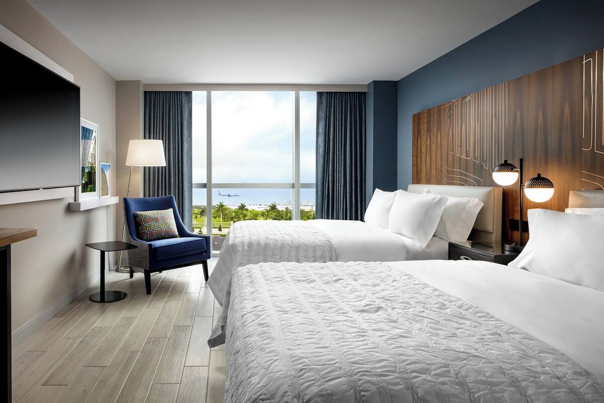 A hotel room with two beds, a TV, a chair, a lamp, and a view of the sky.