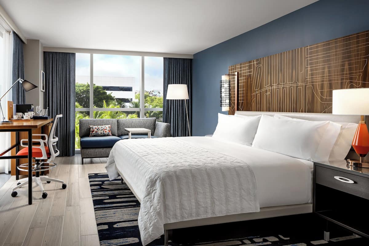 A modern hotel room with a large bed, a desk with a computer, a chair, and a sofa.