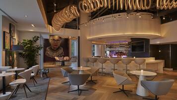A modern bar area with round tables and chairs, a bar counter with bottles, and a large digital screen.
