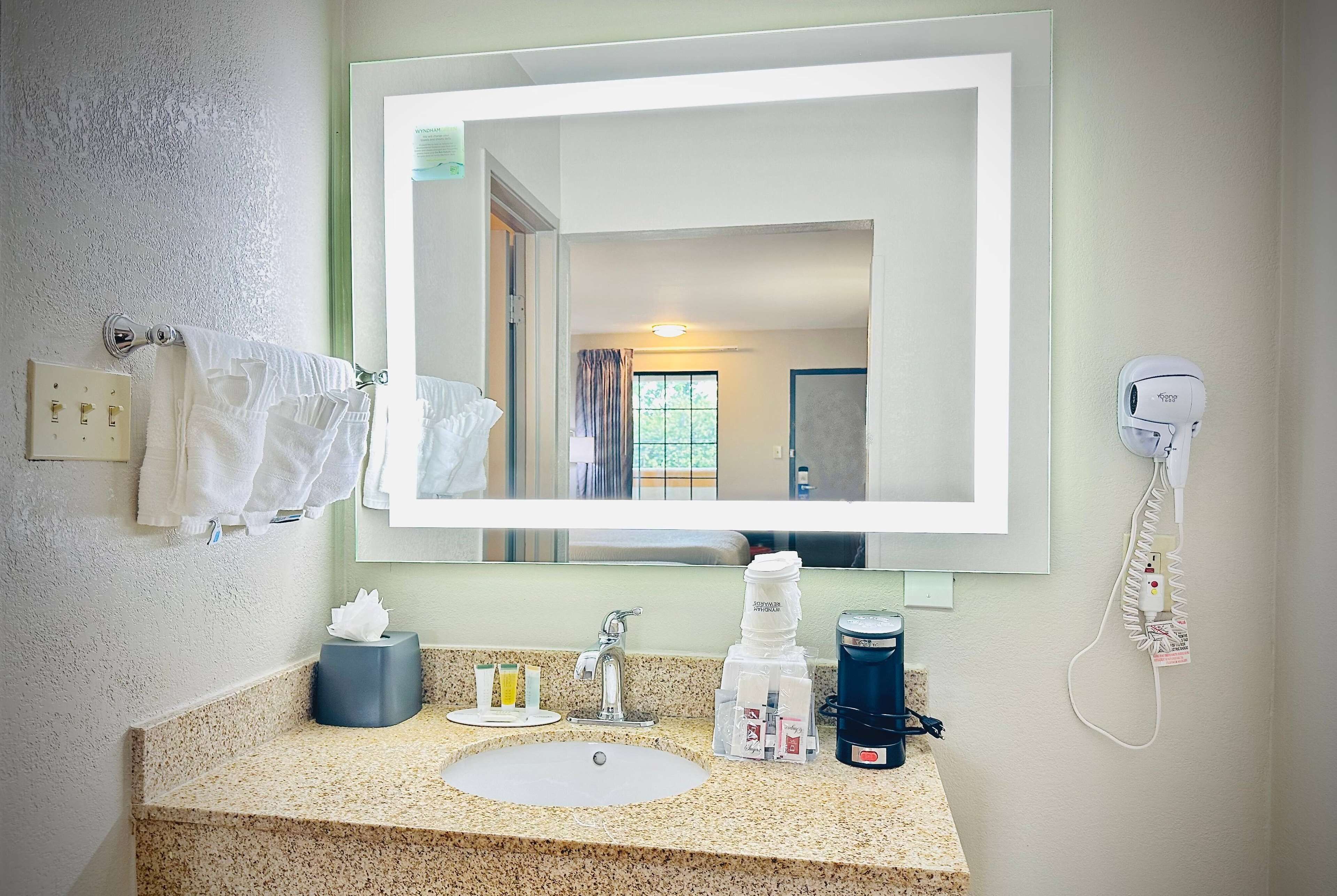 A hotel bathroom with a large mirror, a sink, a towel rack, a hairdryer, and a coffee maker.