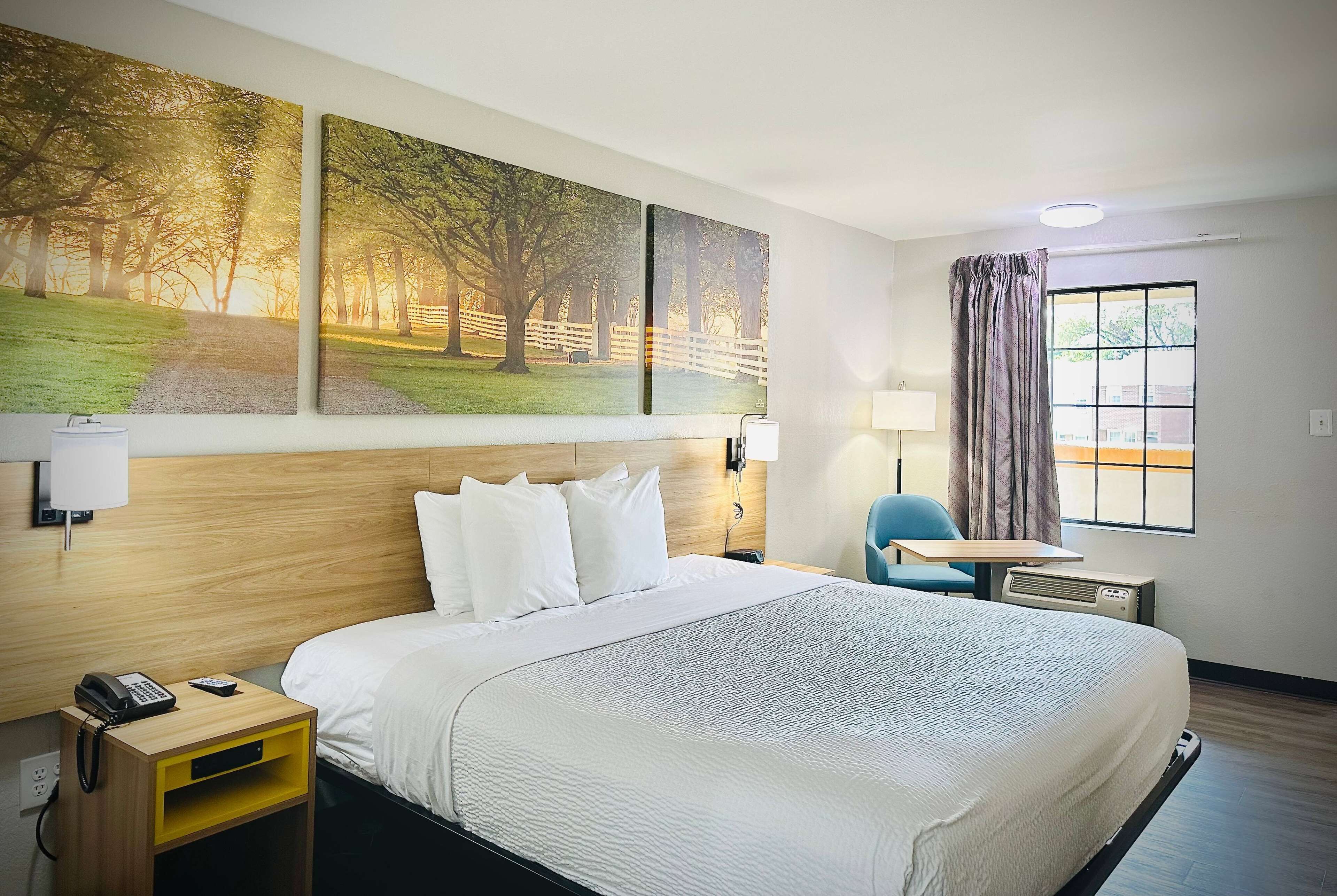 Une chambre d’hôtel avec un grand lit, une table de chevet avec un téléphone, une chaise, une petite table et une fenêtre avec des rideaux.