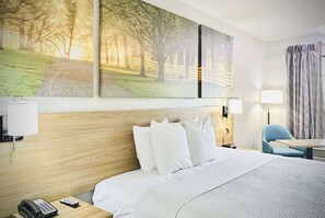 A hotel room with a wooden headboard, white bedding, a desk with a lamp, and a chair.