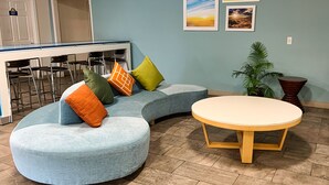 Un espace lounge moderne avec un canapé incurvé, une table ronde et un bar avec des tabourets. Des tableaux encadrés ornent le mur, et une plante en pot est également présente.