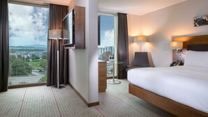 A hotel room with a large bed, a desk, a chair, a TV, and a view of the cityscape. - Hilton Zurich Airport (Opfikon)
