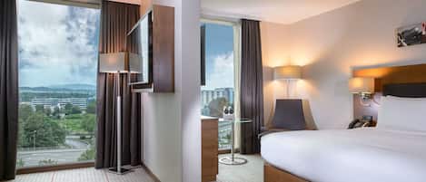 A hotel room with a large bed, a desk, a chair, a TV, and a view of the cityscape.