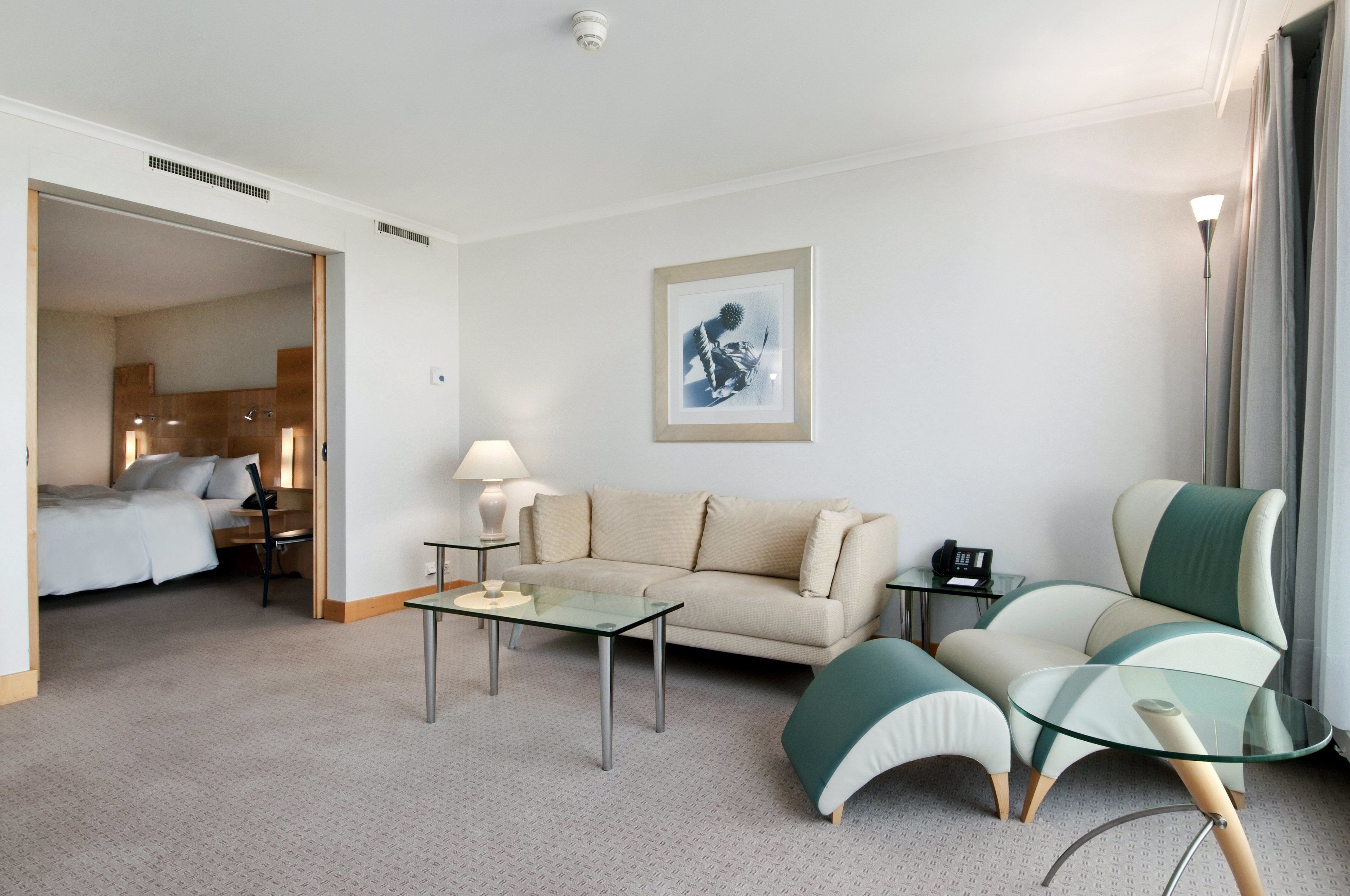Une chambre d’hôtel avec un canapé, une table basse en verre, une lampe, une œuvre d’art encadrée au mur et un lit à l’arrière-plan.