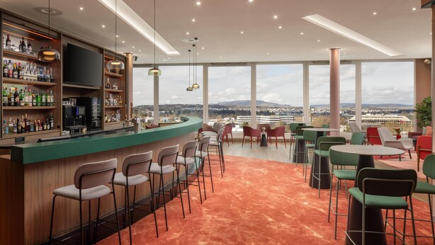 A bar area with a view of the cityscape, bar stools, and a mix of red and green chairs. - Hilton Zurich Airport (Opfikon)