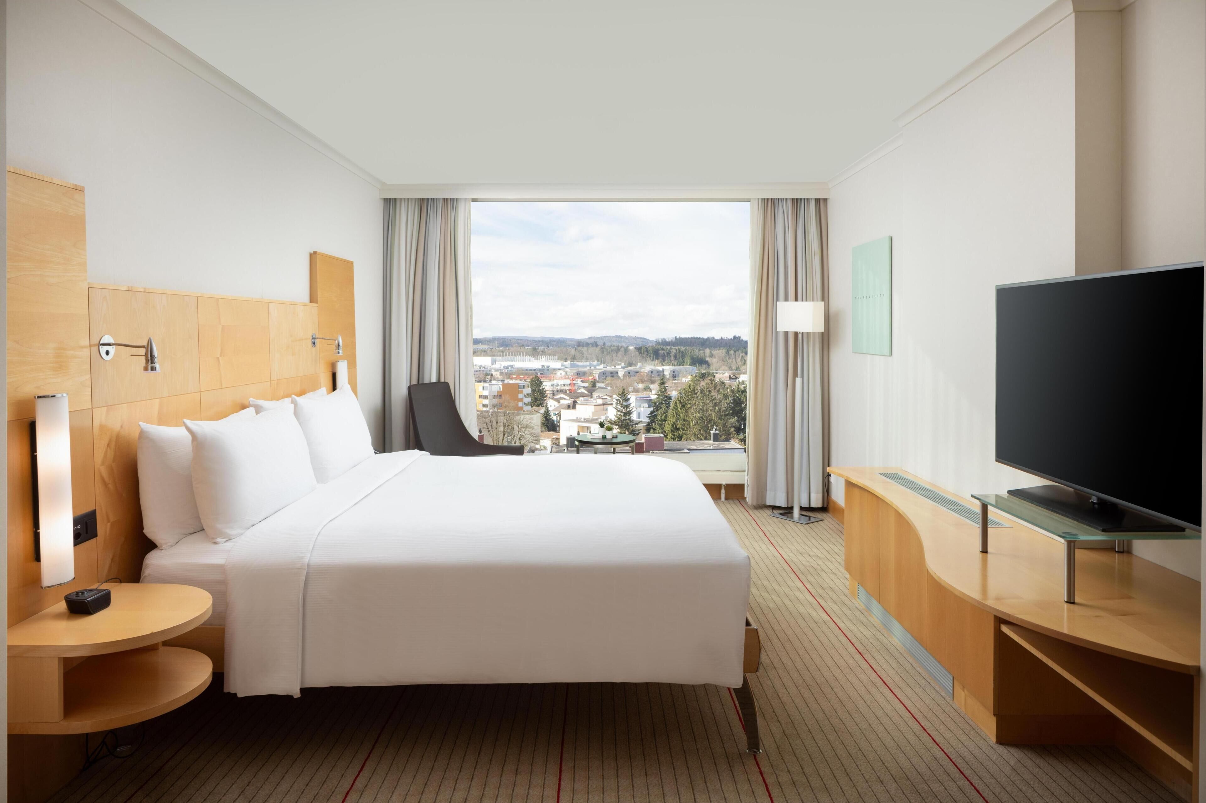 A hotel room with a large bed, a TV, a desk, and a view of a cityscape.
