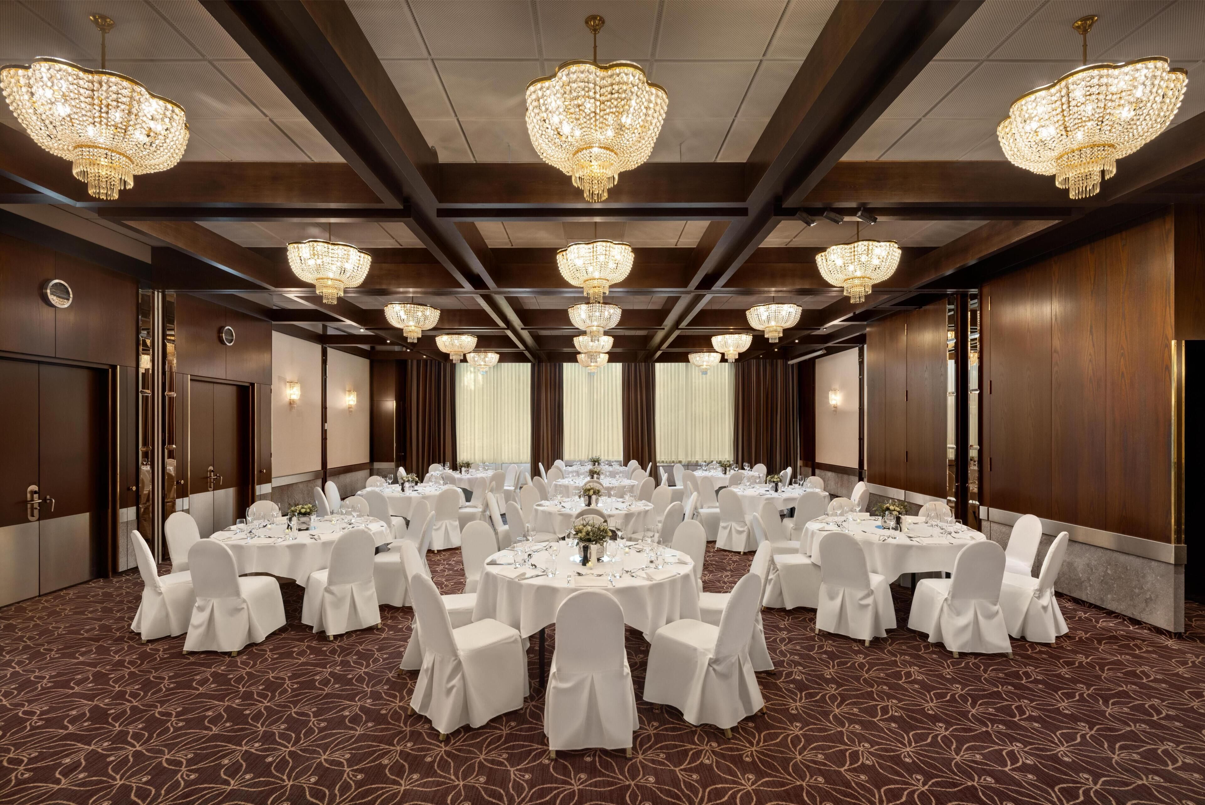A banquet hall with round tables, white chairs, chandeliers, and wooden paneling.