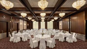 A banquet hall with round tables, white chairs, chandeliers, and wooden paneling.