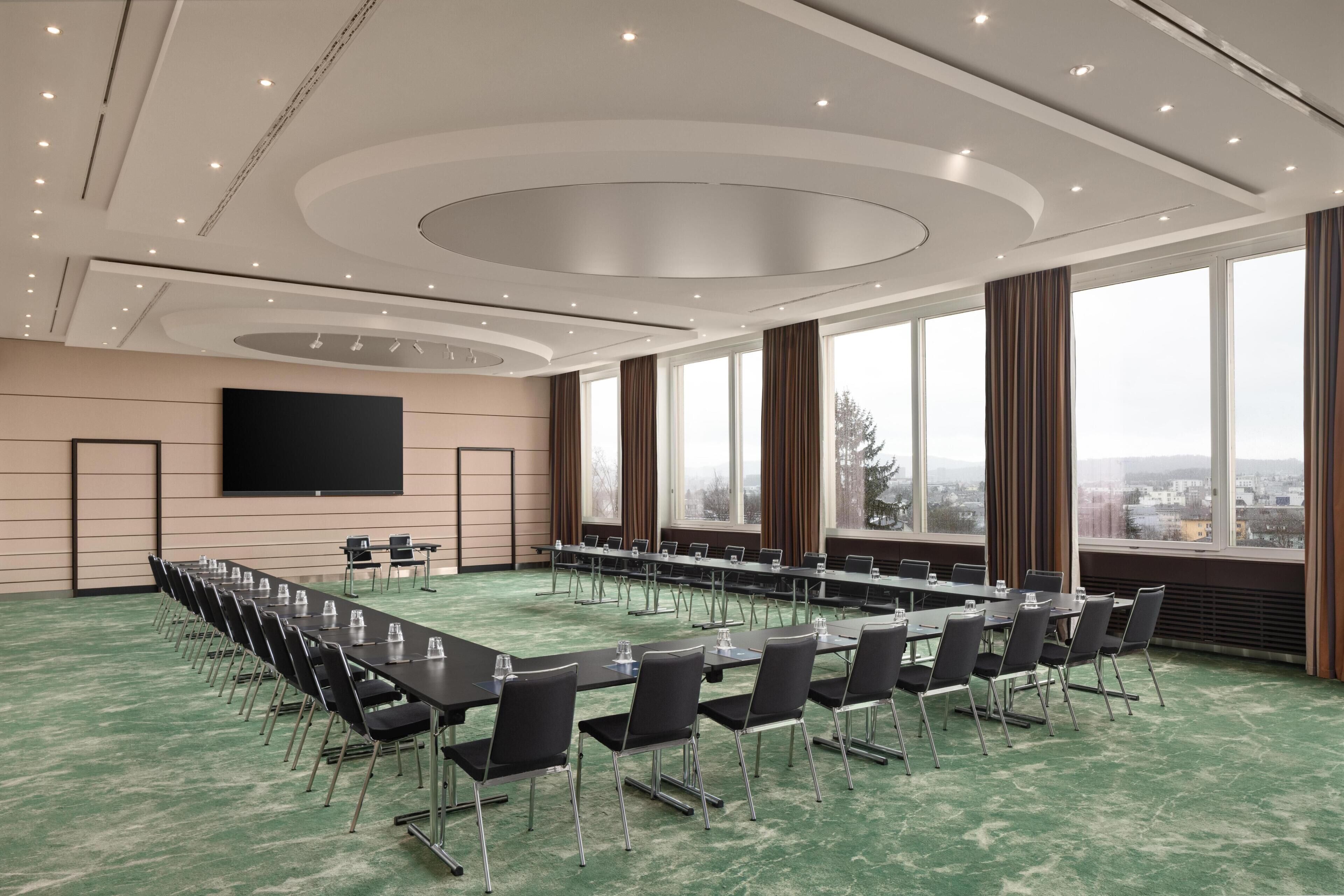 A conference room with a long table, chairs, a flat-screen TV, and large windows with a city view.