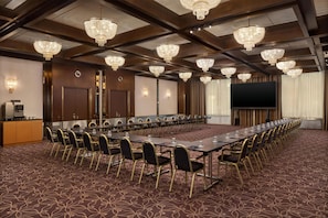 A conference room with a long table, chairs, chandeliers, a projector screen, and a coffee station.