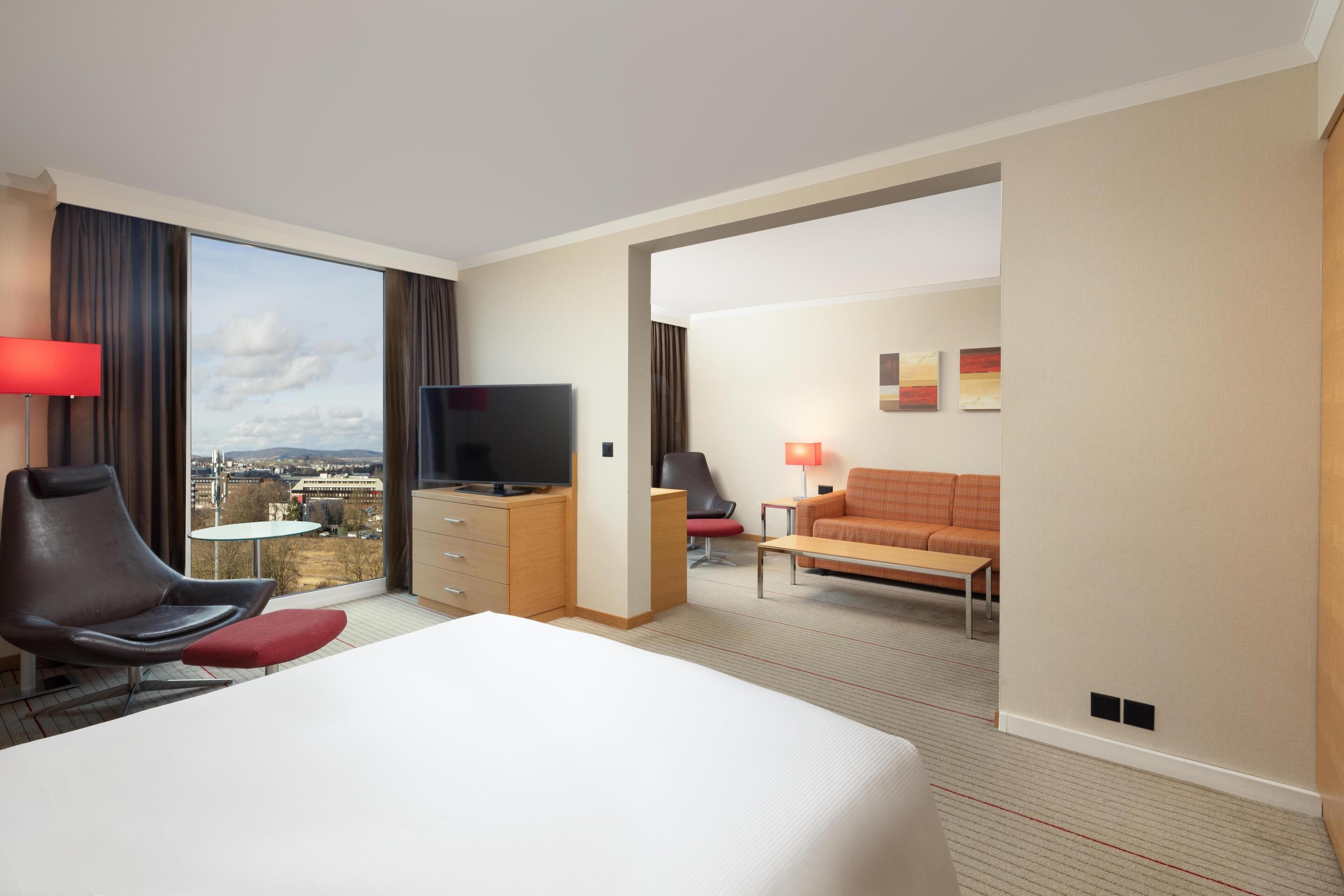 A hotel room with a bed, a TV, a desk, a chair, a sofa, and a view of the city.