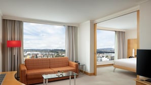 A hotel room with a sofa, a glass coffee table, a bed, a TV, and a city view. - Hilton Zurich Airport (Opfikon)