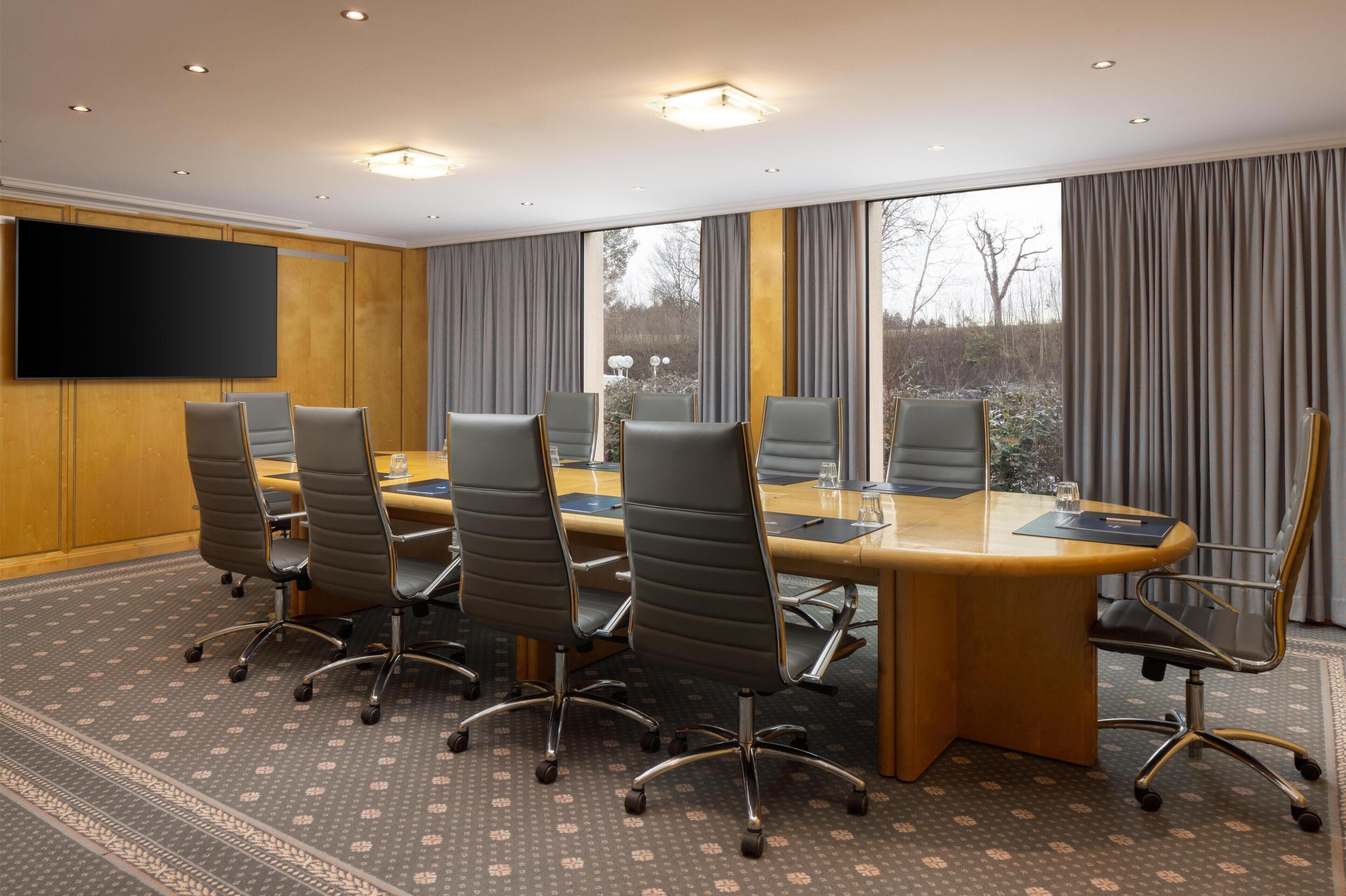 A conference room with a large table, six chairs, a flat-screen TV, and large windows with curtains.